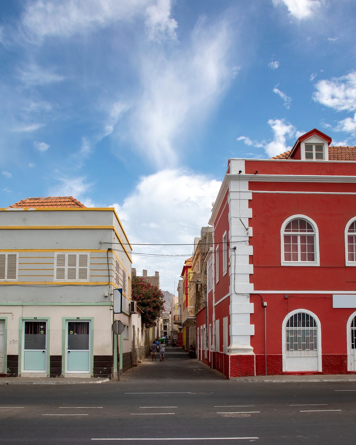 Street view featuring a vivid red colonial-style building on the right and a pale blue and yellow building on the left, with a narrow alley between them, a few pedestrians in the distance and soft clouds drifting across a bright blue sky.