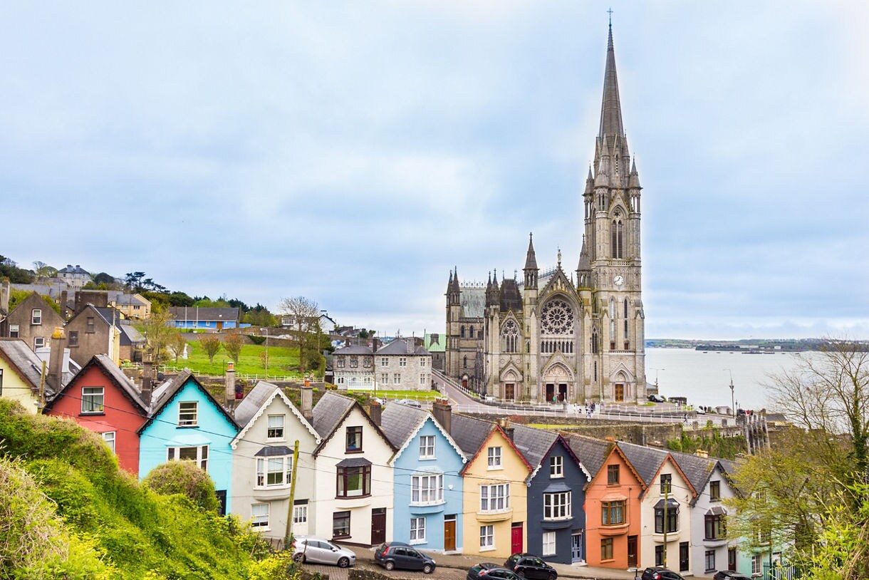 Colorful row of houses on a hill in Cobh, Ireland, with the large Gothic-style St. Colman’s Cathedral and its tall spire overlooking the sea in the background.