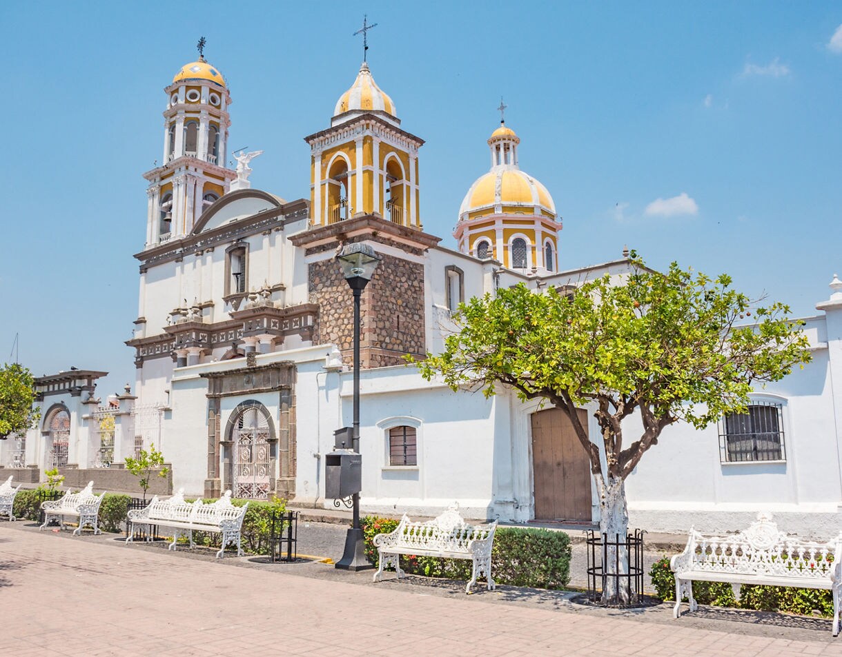 White colonial church in Comala with yellow domes and twin bell towers, surrounded by benches and trees along the plaza.