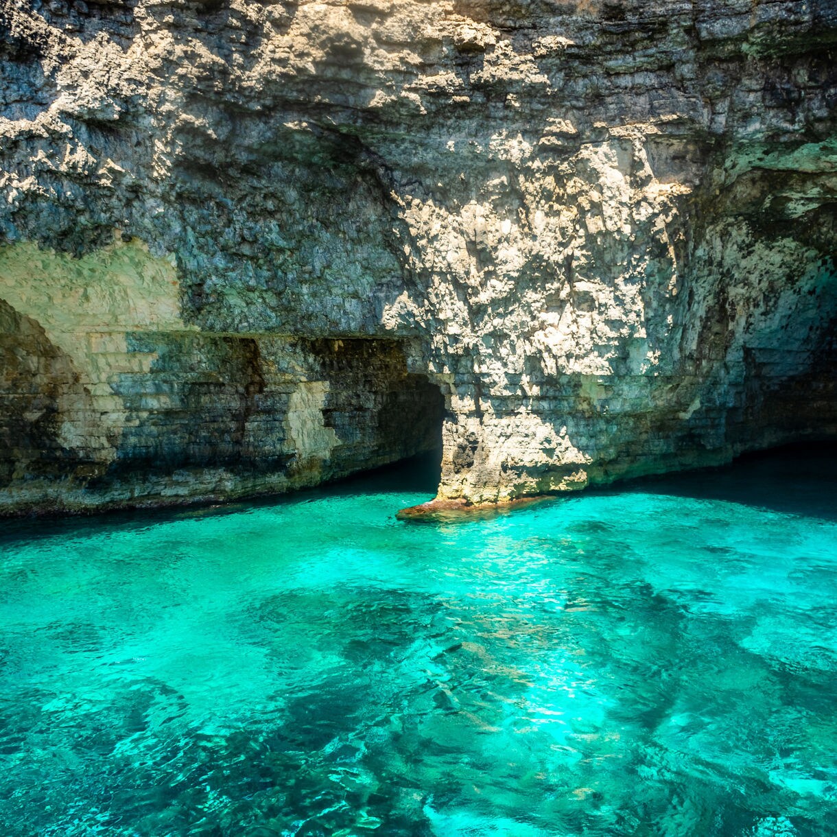 Rocky sea cave on Comino Island, Malta, with clear turquoise water reflecting light onto limestone walls.