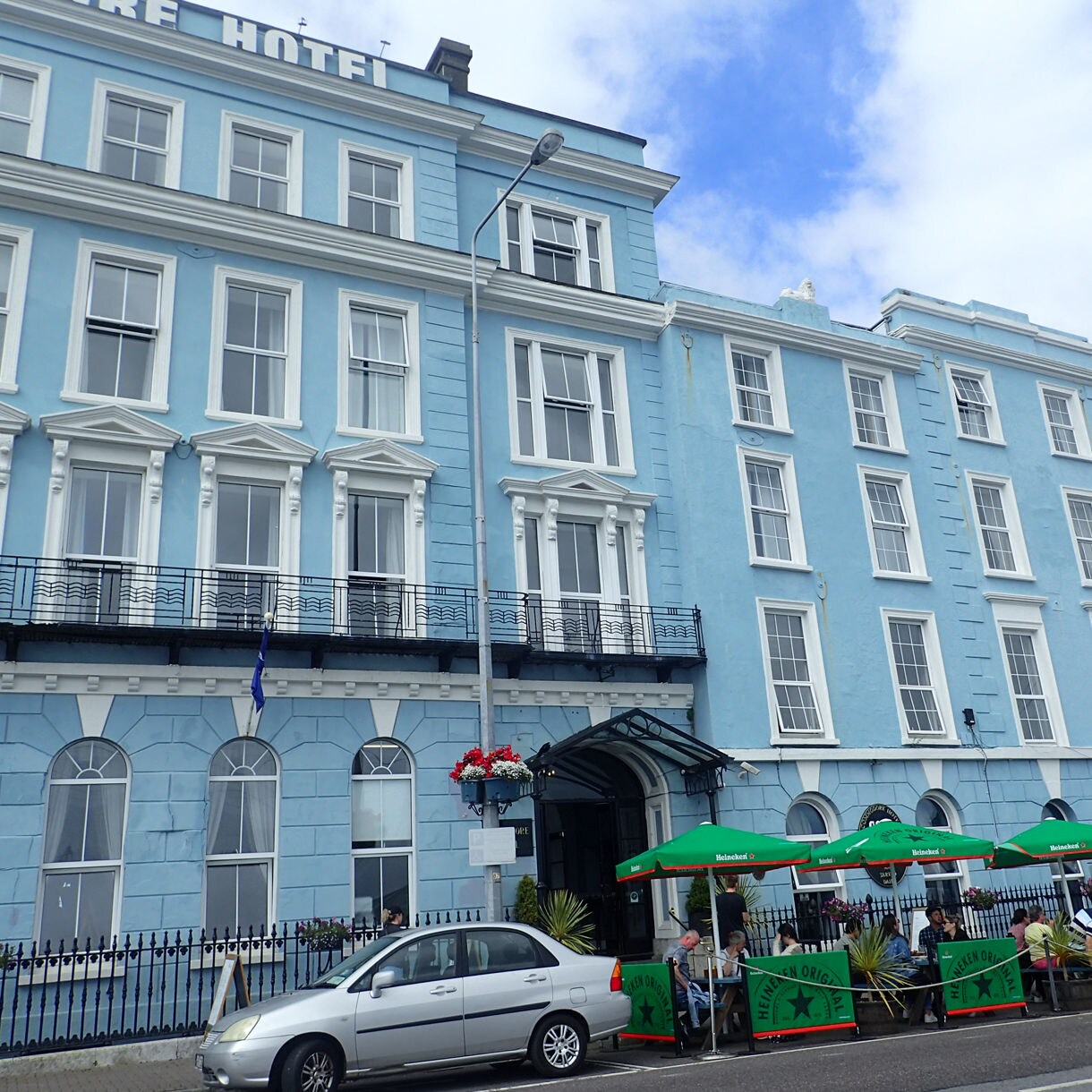 Bright blue exterior of the Commodore Hotel in Cobh, Ireland, with green umbrellas shading outdoor seating where people dine along the sidewalk.