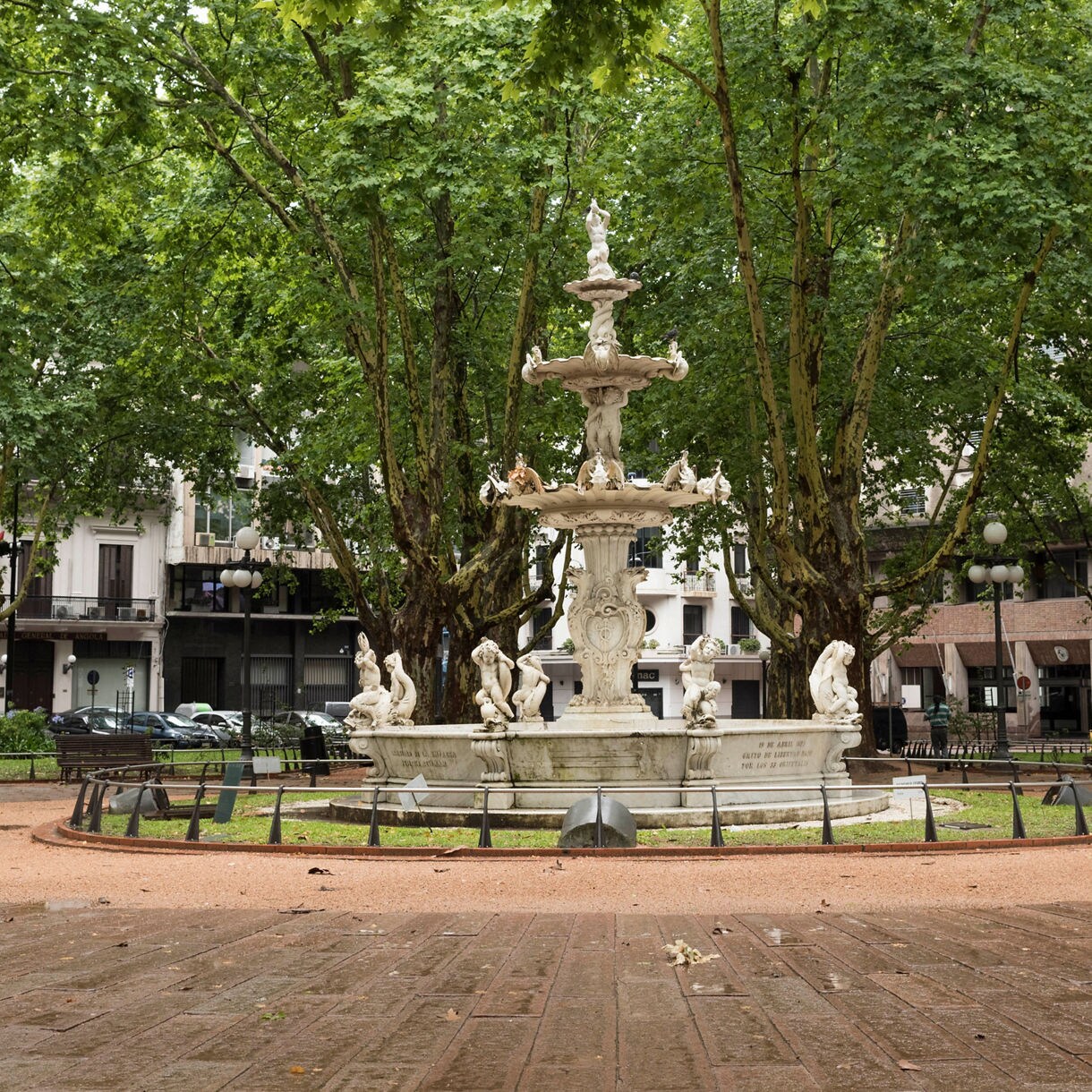 Ornate white marble fountain with sculpted figures in the center of a tree-lined plaza surrounded by benches and historic buildings.
