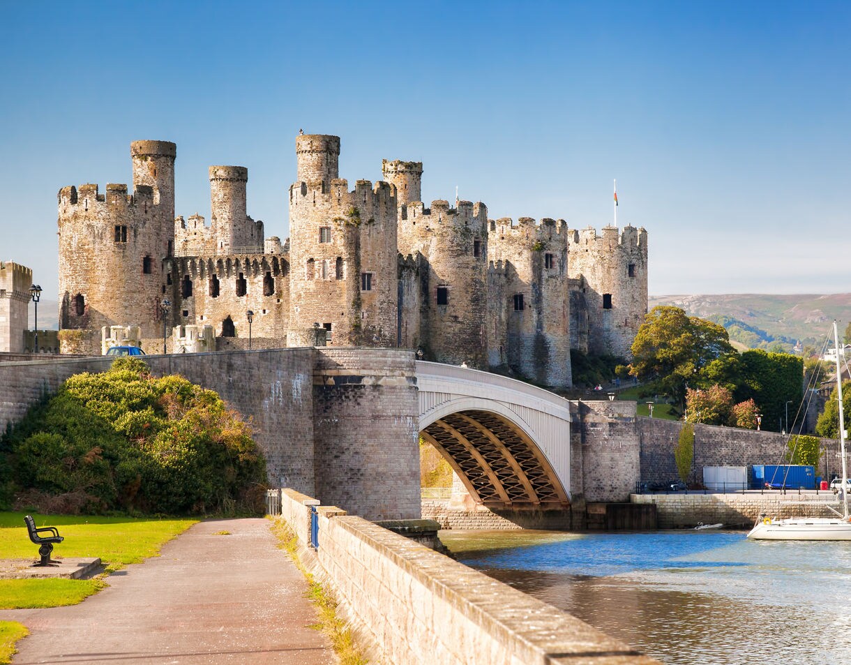 Conwy Castle rises above the river in Wales, with stone towers, a sweeping bridge and sailboats drifting under a clear blue sky