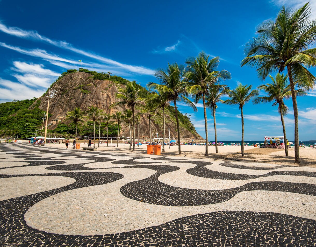 Copacabana Beach promenade with its black and white wave-pattern mosaic, tall palm trees, golden sand and a rocky green hill rising in the background under a bright blue sky.