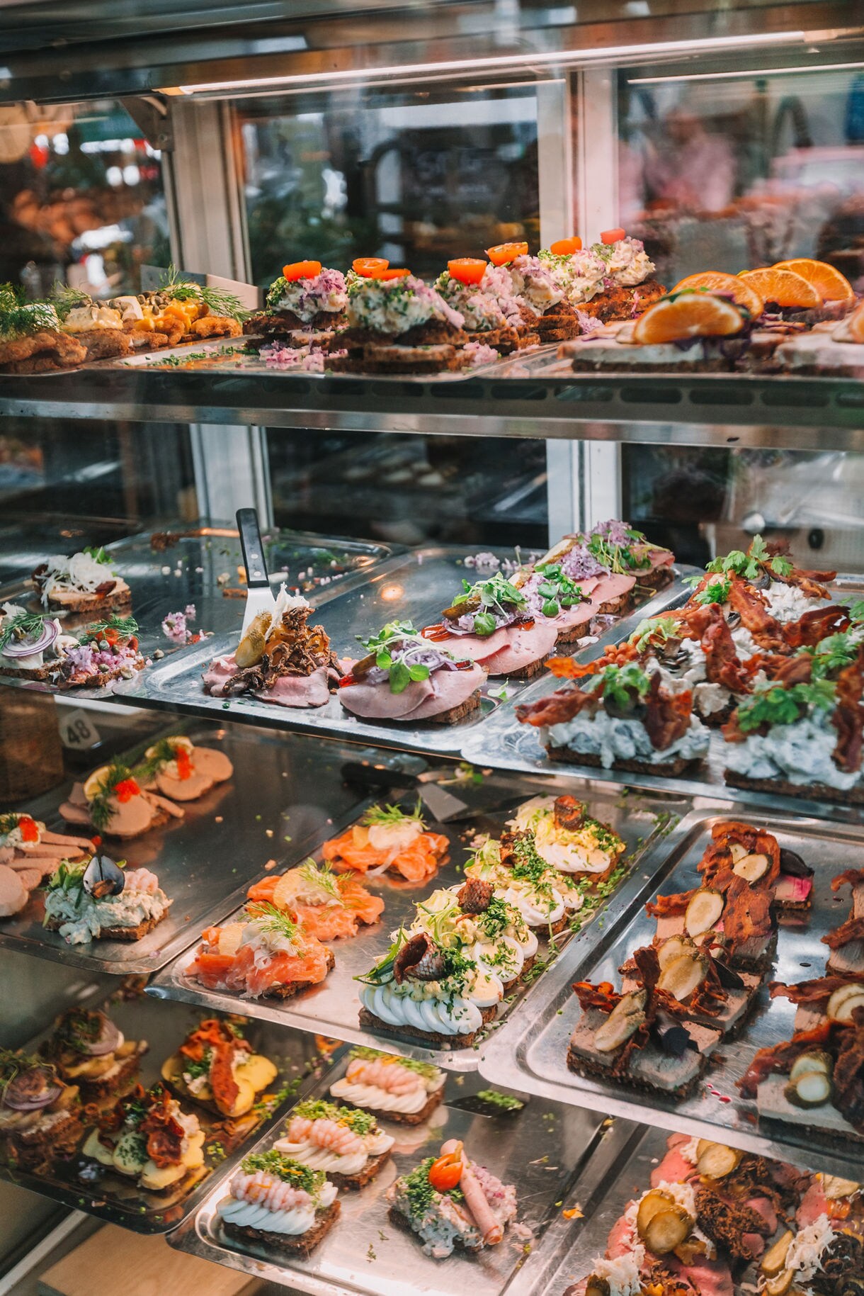 Display case filled with colorful Danish open-faced sandwiches topped with fish, meats, and herbs.