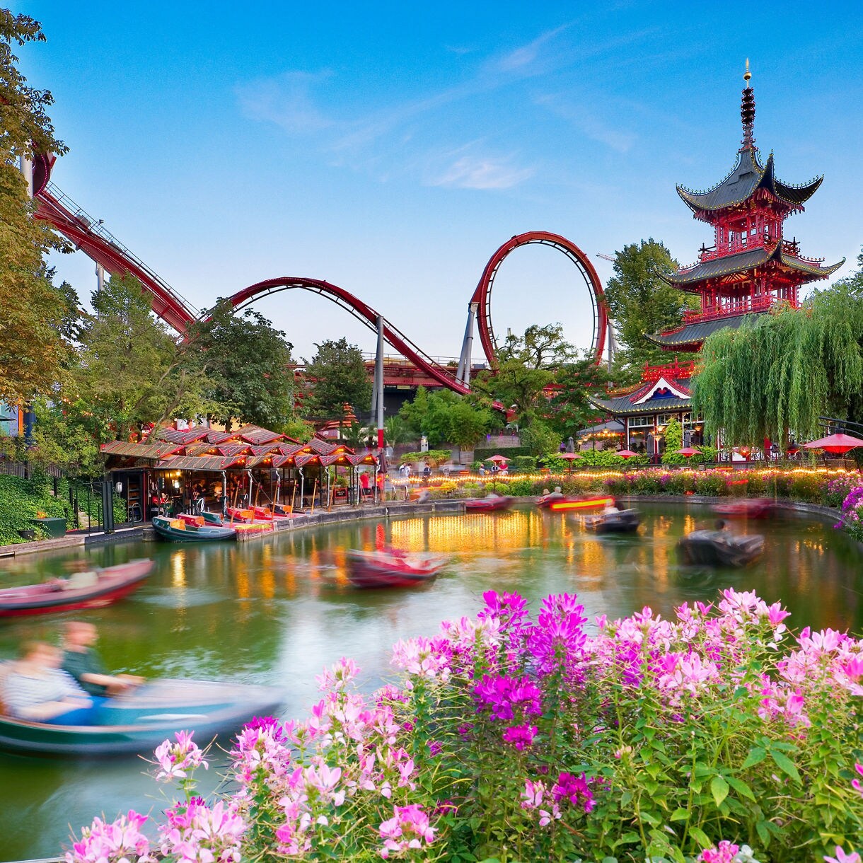 Tivoli Gardens in Copenhagen with colorful flowers, boats on a canal, a red pagoda, and roller coaster loops.