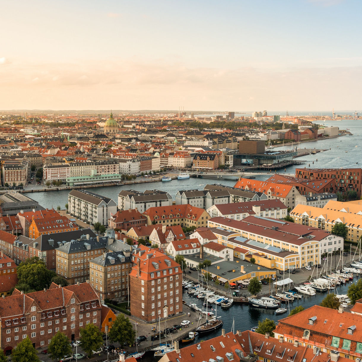 Wide panoramic aerial view of Copenhagen at sunset, showing red-roofed buildings, canals, docked boats and the city skyline in the distance.
