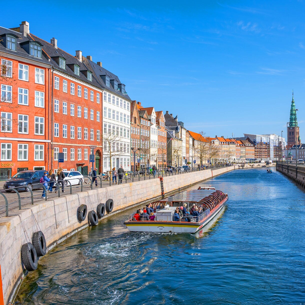 Sightseeing boat gliding along a Copenhagen canal lined with historic buildings and church spires.