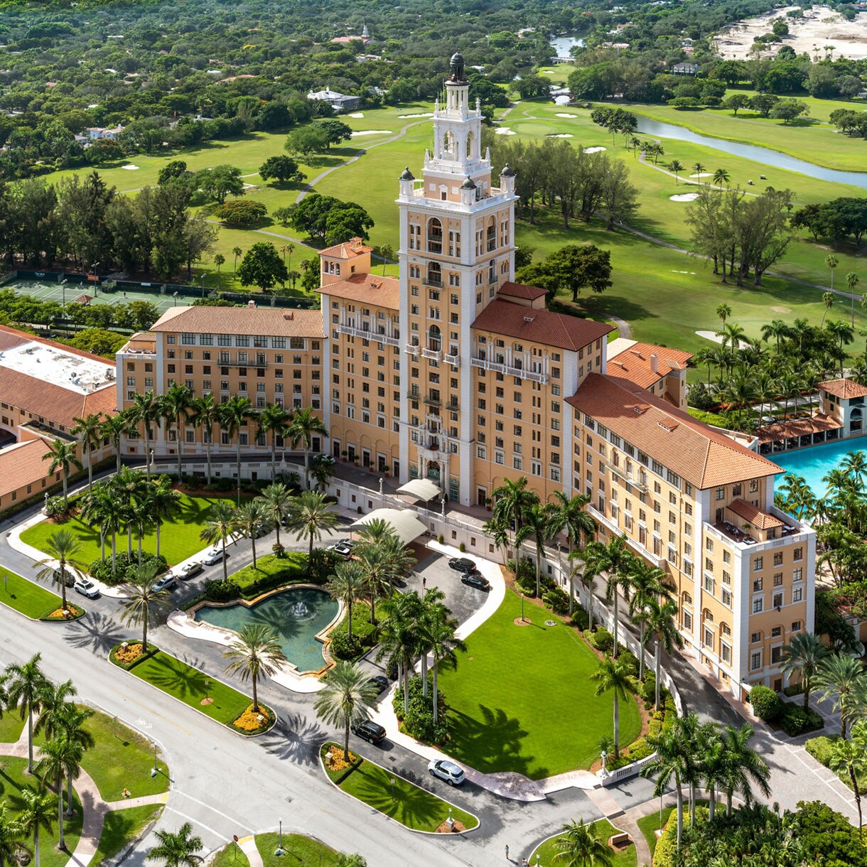 Aerial view of the Biltmore Hotel in Coral Gables, Florida, with its tall central tower, red-tiled roofs, surrounding gardens, pool and nearby golf course.