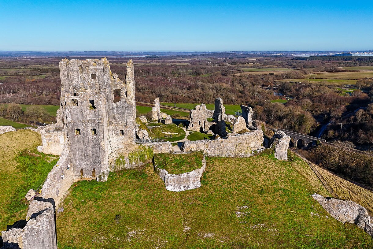 Aerial view of Corfe Castle ruins on a grassy hilltop in Dorset, with crumbling stone walls and towers surrounded by countryside and distant woodlands.