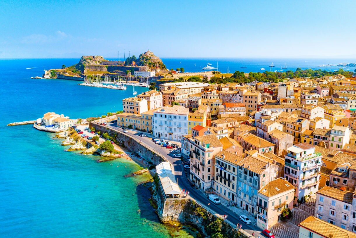 Aerial view of Corfu, Greece, showing pastel-colored houses, a seaside fortress, a marina, and the bright turquoise waters of the Ionian Sea.