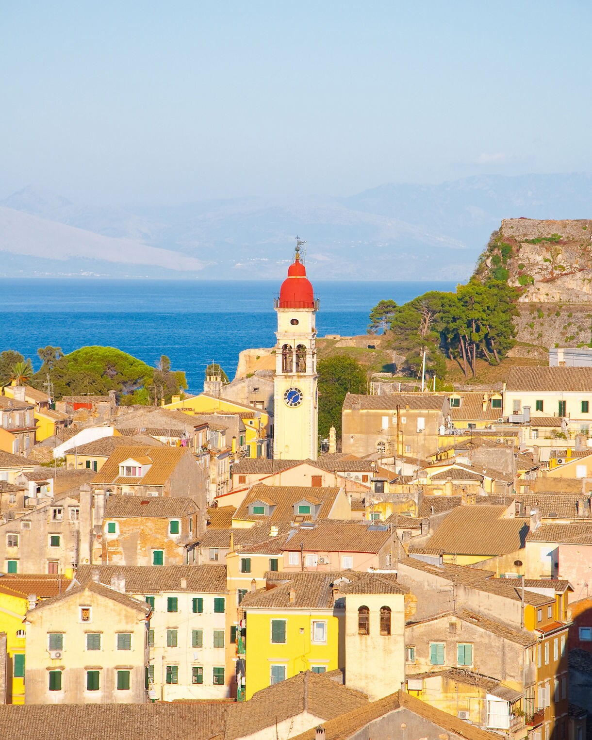Aerial view of Corfu’s old town with stone houses and the tall red-domed bell tower of St. Spyridon Church set against the blue sea and distant mountains.