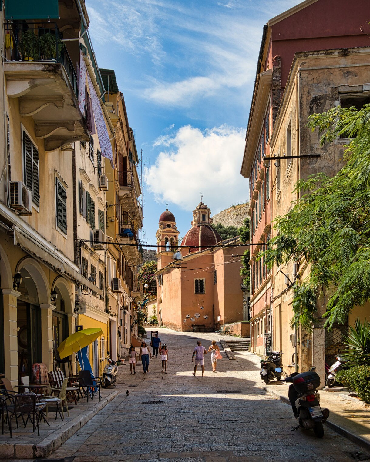 Narrow cobblestone street in Corfu lined with colorful old buildings, leading to a church with red domes under a bright blue sky.