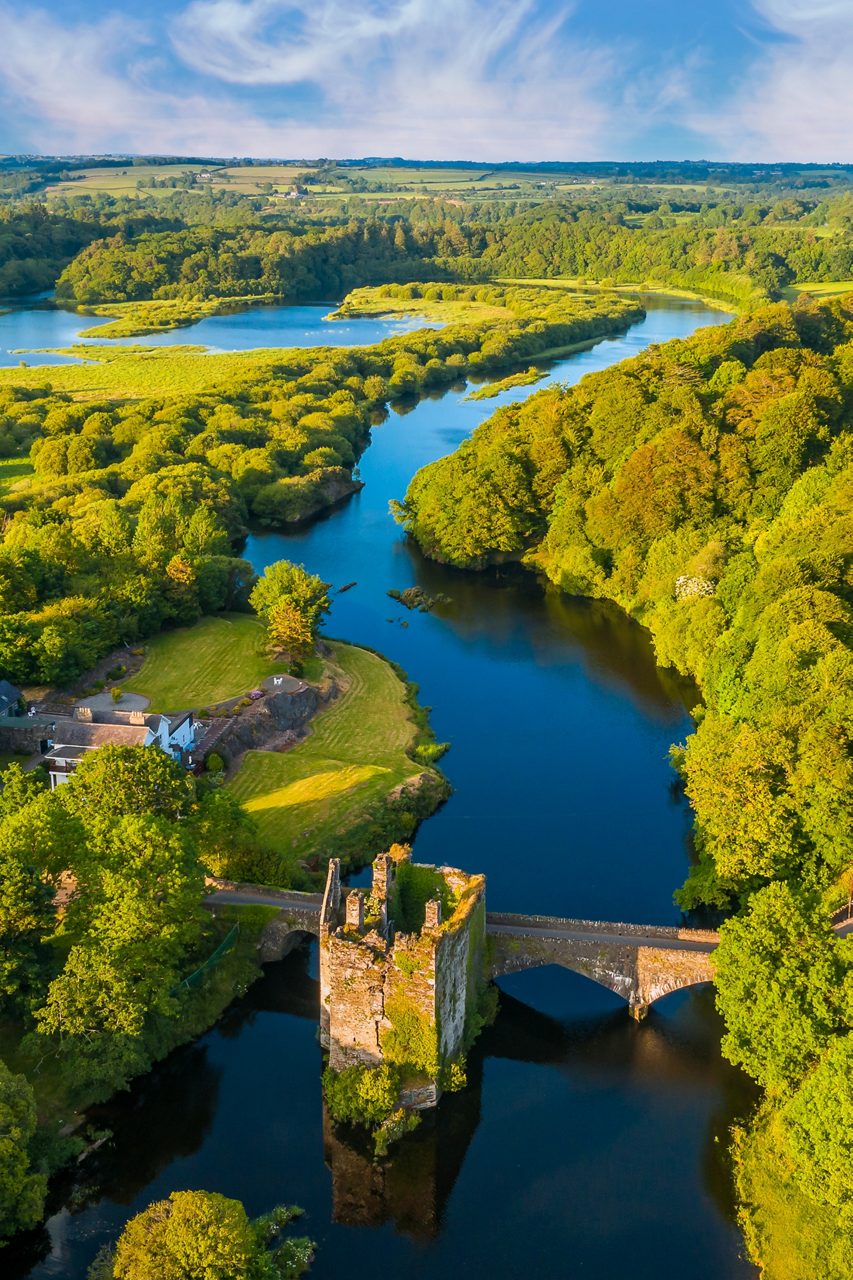 Aerial view of a winding river bordered by dense green trees with a small stone bridge and castle ruins in the foreground.