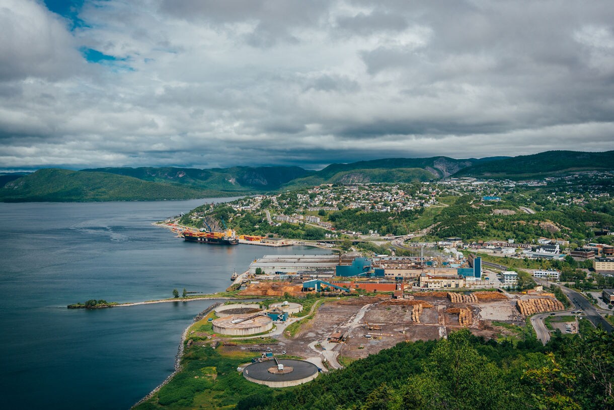 A wide aerial view of Corner Brook, Newfoundland, showing a deep blue bay, industrial waterfront, stacked timber yards, residential hillsides and surrounding green mountains under a cloudy sky.
