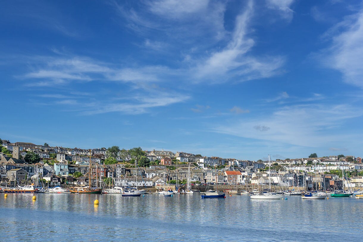 View of Falmouth harbor in Cornwall with sailboats and yachts moored in calm water, backed by hillside houses under a blue sky.