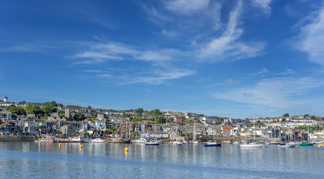 View of Falmouth harbor in Cornwall with sailboats and yachts moored in calm water, backed by hillside houses under a blue sky.