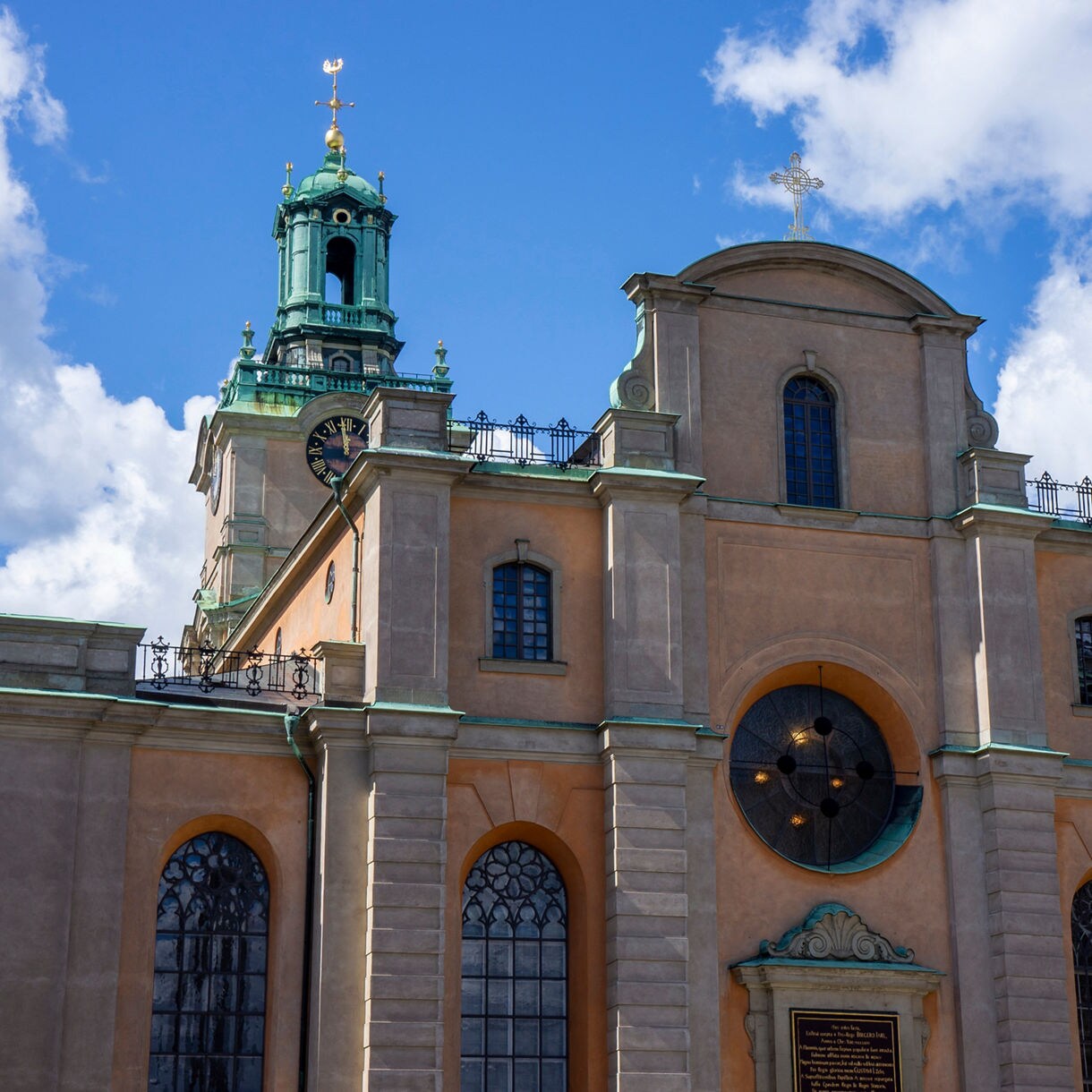 Exterior of Stockholm Cathedral, also known as Storkyrkan, featuring tall arched windows, a clock tower with a green copper roof and ornate details under a bright blue sky.