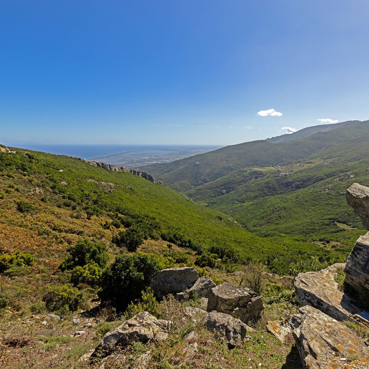 View over Corsica’s inland mountains and valleys, with rocky outcrops and green hills descending toward the distant blue sea.