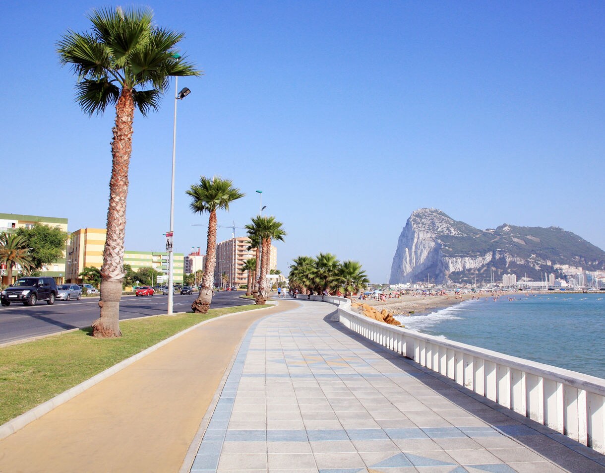 A seaside promenade lined with palm trees beside a sandy beach, with the Rock of Gibraltar visible across the water under a clear blue sky.