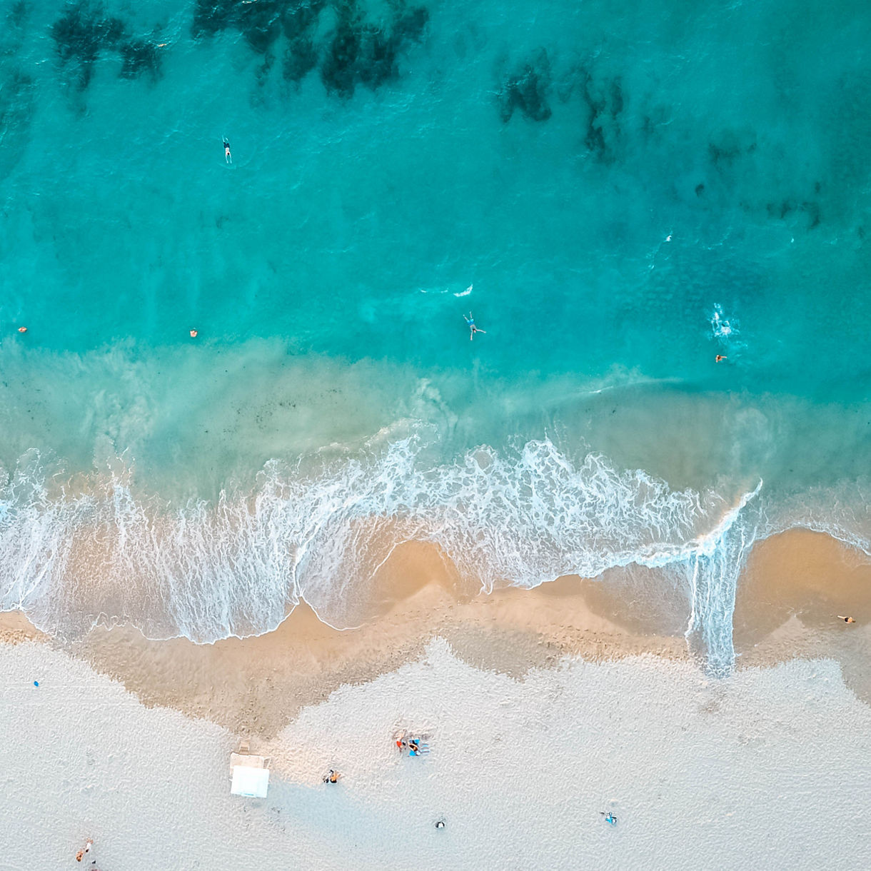 Aerial view of Cottesloe Beach with turquoise waves rolling onto golden sand, swimmers in the water and people relaxing onshore.
