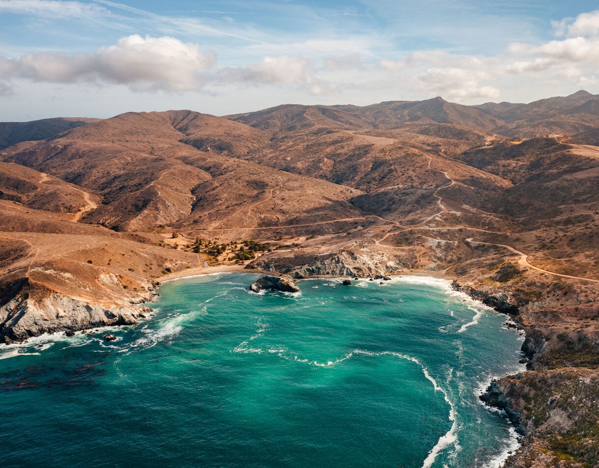 Aerial view of a secluded cove on Catalina Island with bright turquoise water, rocky cliffs and winding dirt roads stretching across dry, rolling hills.