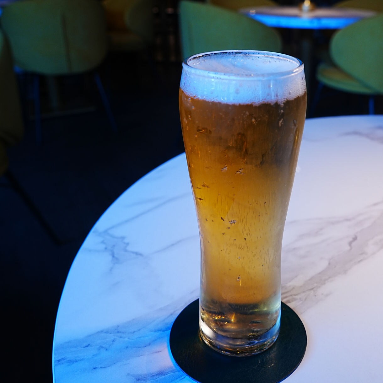 Tall chilled beer glass on a marble table, light foam at the top, with another beer in the background.