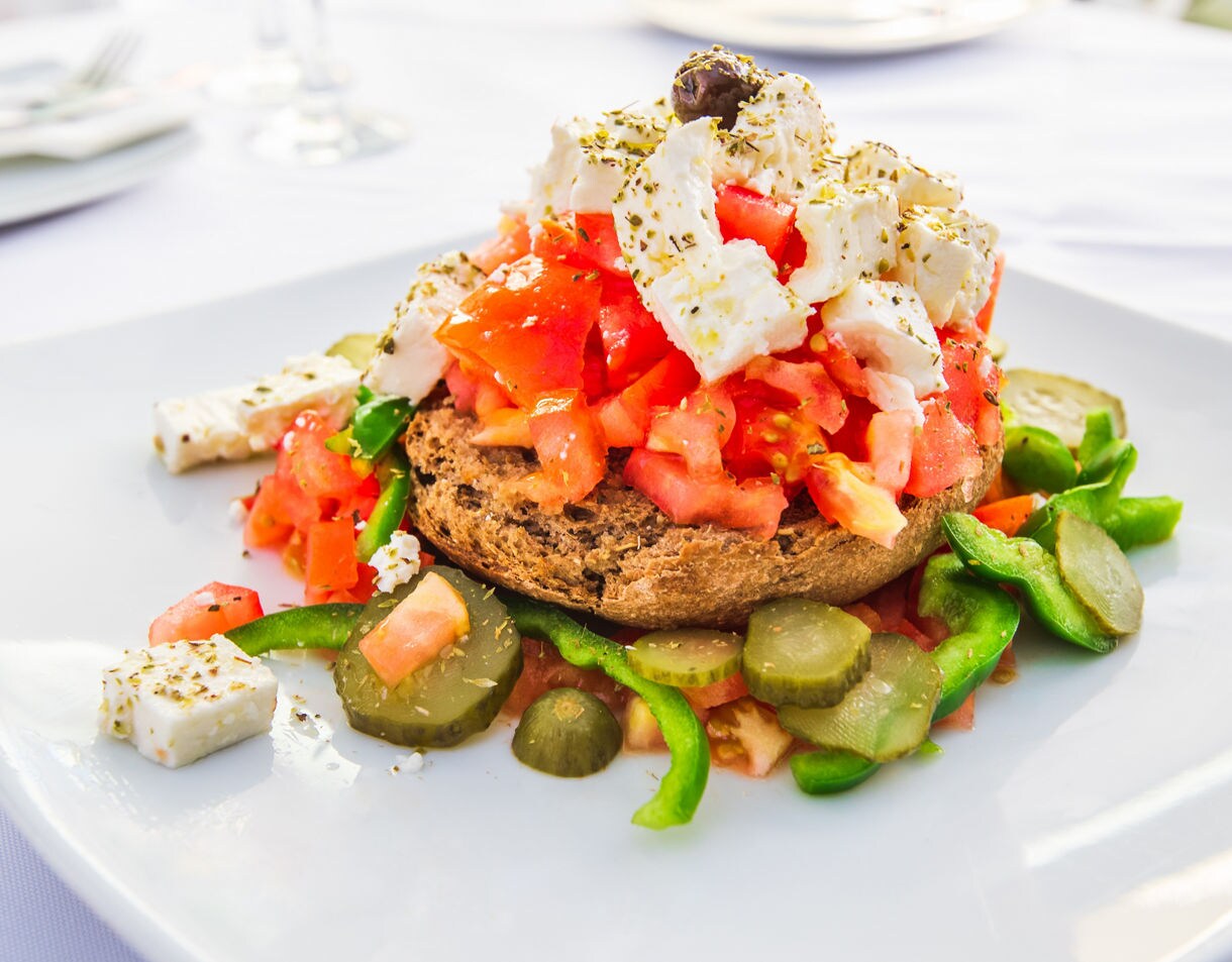 Close-up of a Cretan dakos salad featuring chopped tomatoes, feta cheese, green peppers and pickles on a barley rusk, served on a white plate.