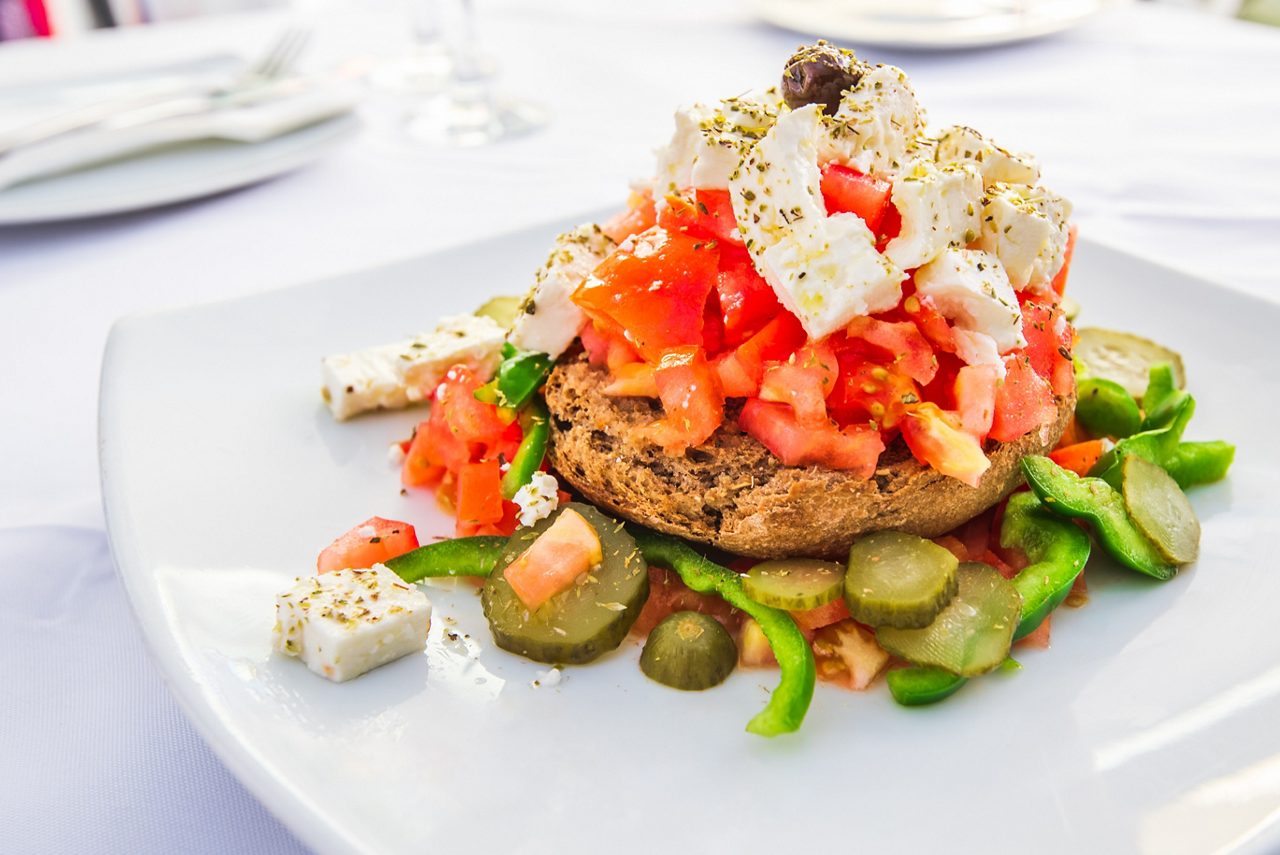 A white plate bearing a Greek-style open-faced salad with diced tomatoes, crumbled feta cheese, olives, sliced cucumbers, and green peppers piled atop a round piece of rustic bread, garnished with dried herbs.