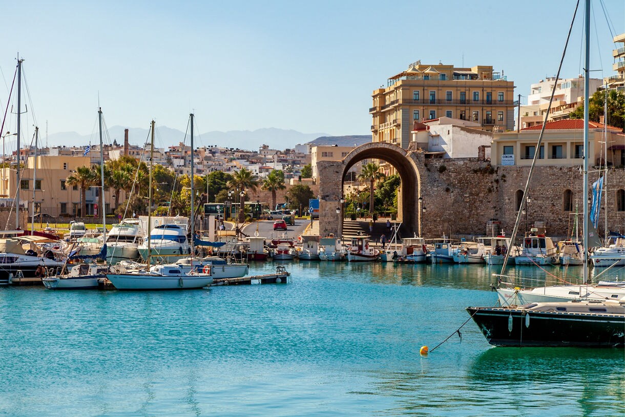 View of Heraklion’s marina with docked sailboats, a stone archway, waterfront buildings and a backdrop of distant mountains under a clear sky.