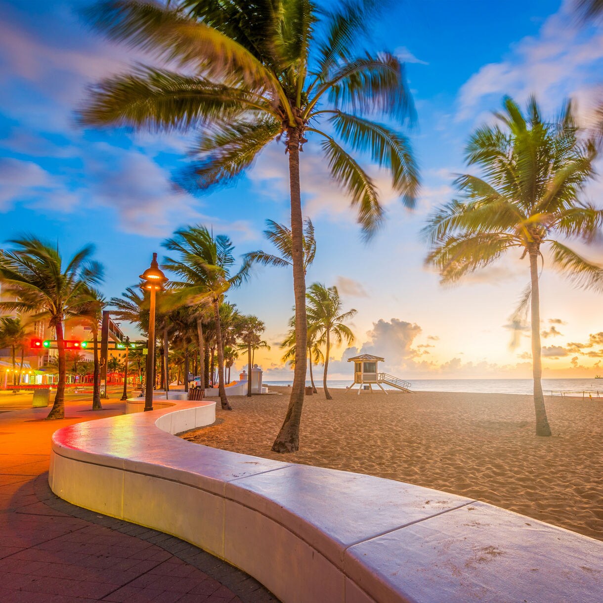 Palm trees along Fort Lauderdale Beach at sunrise with a lifeguard tower, sandy shoreline and colorful sky reflecting on the boardwalk.