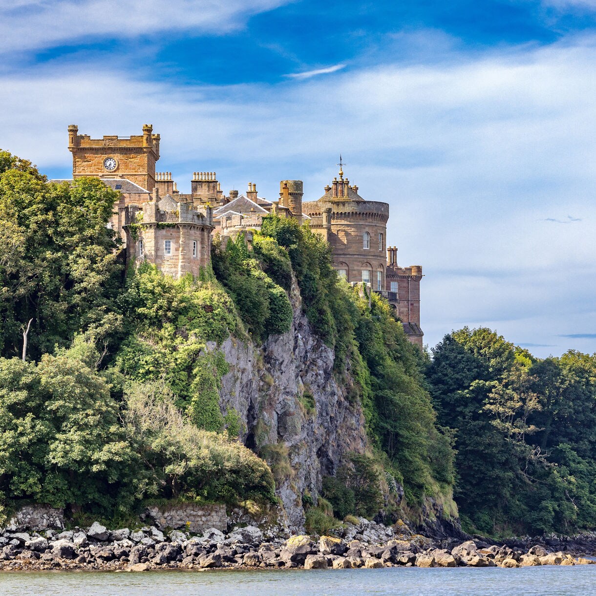 Stone towers and walls of Culzean Castle rising above a steep, rocky cliff covered in greenery, overlooking the shoreline and water under a blue sky.