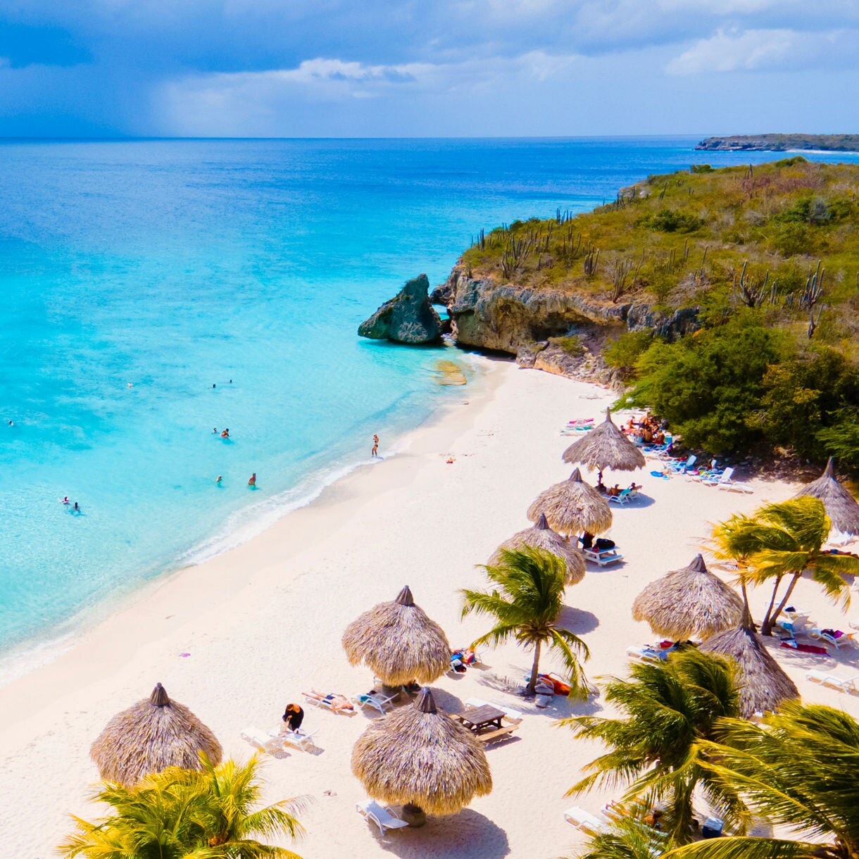 Aerial view of a white sand beach lined with thatched umbrellas and palm trees along turquoise water in a rocky green bay in Curaçao.