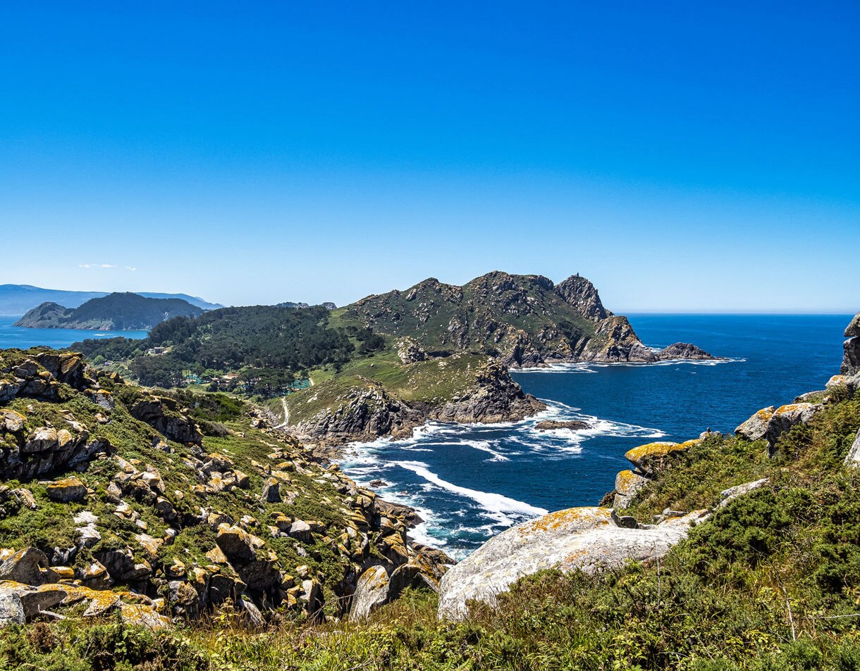 Rocky coastal cliffs of the Cíes Islands with deep blue ocean waves and green hills under a clear blue sky.