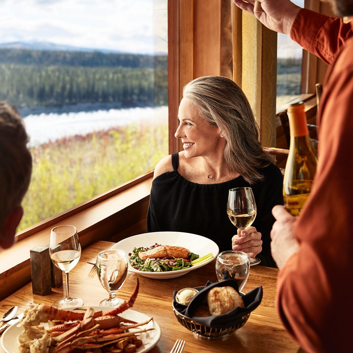 Two women enjoying coffee together at a coffee bar counter, with one wearing a striped top and the other in a yellow sweater, pastry display case visible beside them.