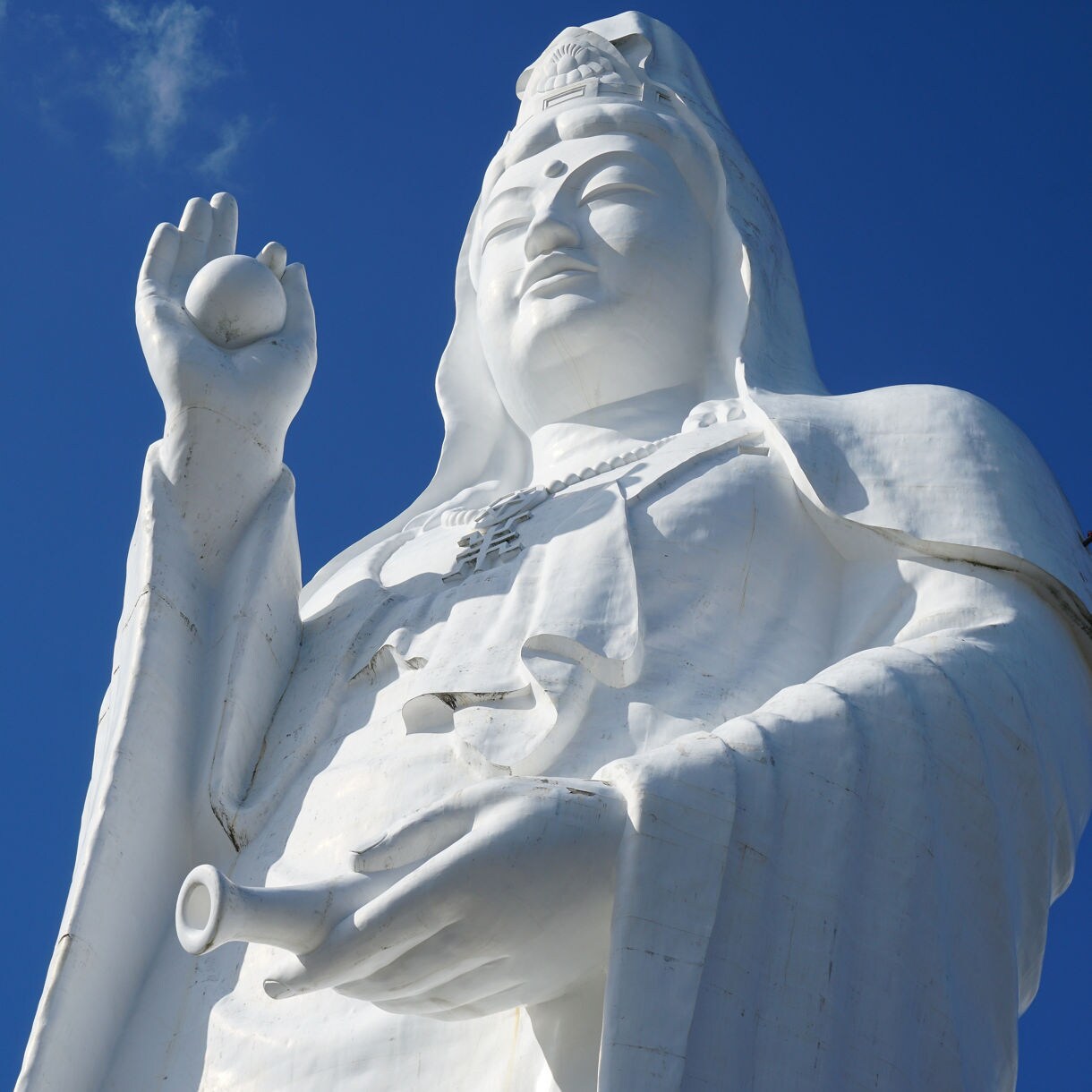 Low-angle view of a massive white Daikannon statue holding a round object in one hand and a small vessel in the other, set against a bright blue sky.