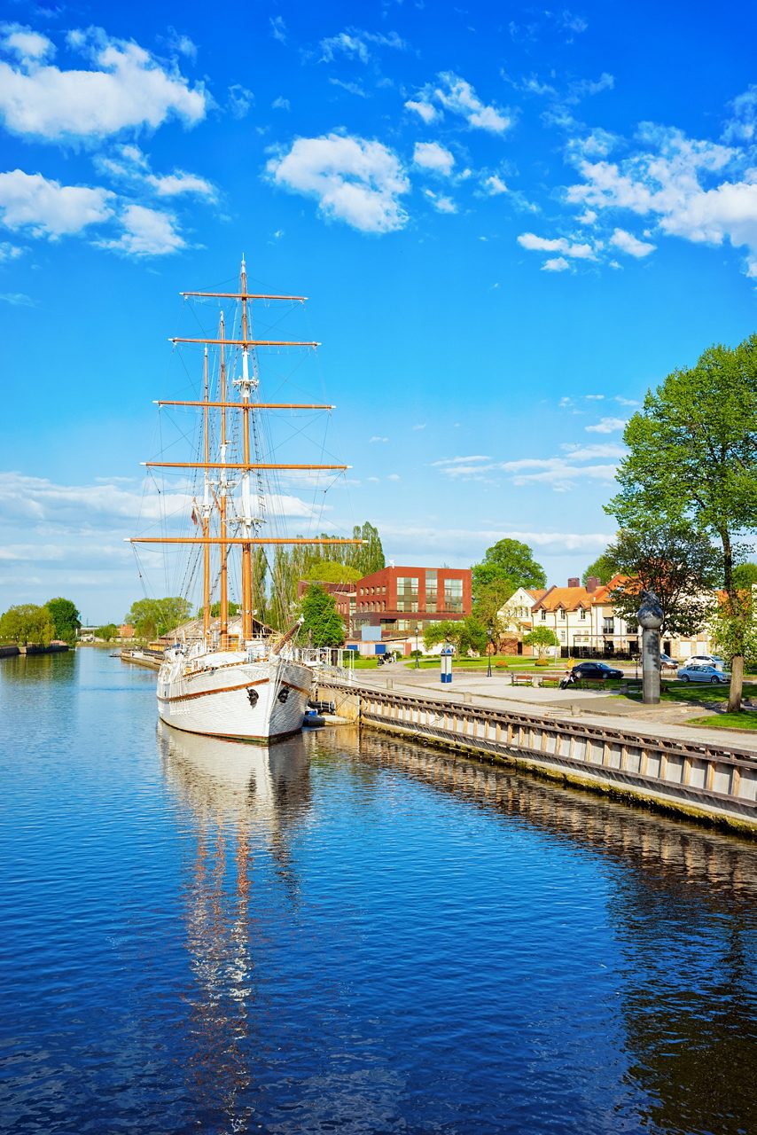 White sailing ship moored on the Dane River in Klaipėda, Lithuania, reflecting on the water with old town buildings in the background.