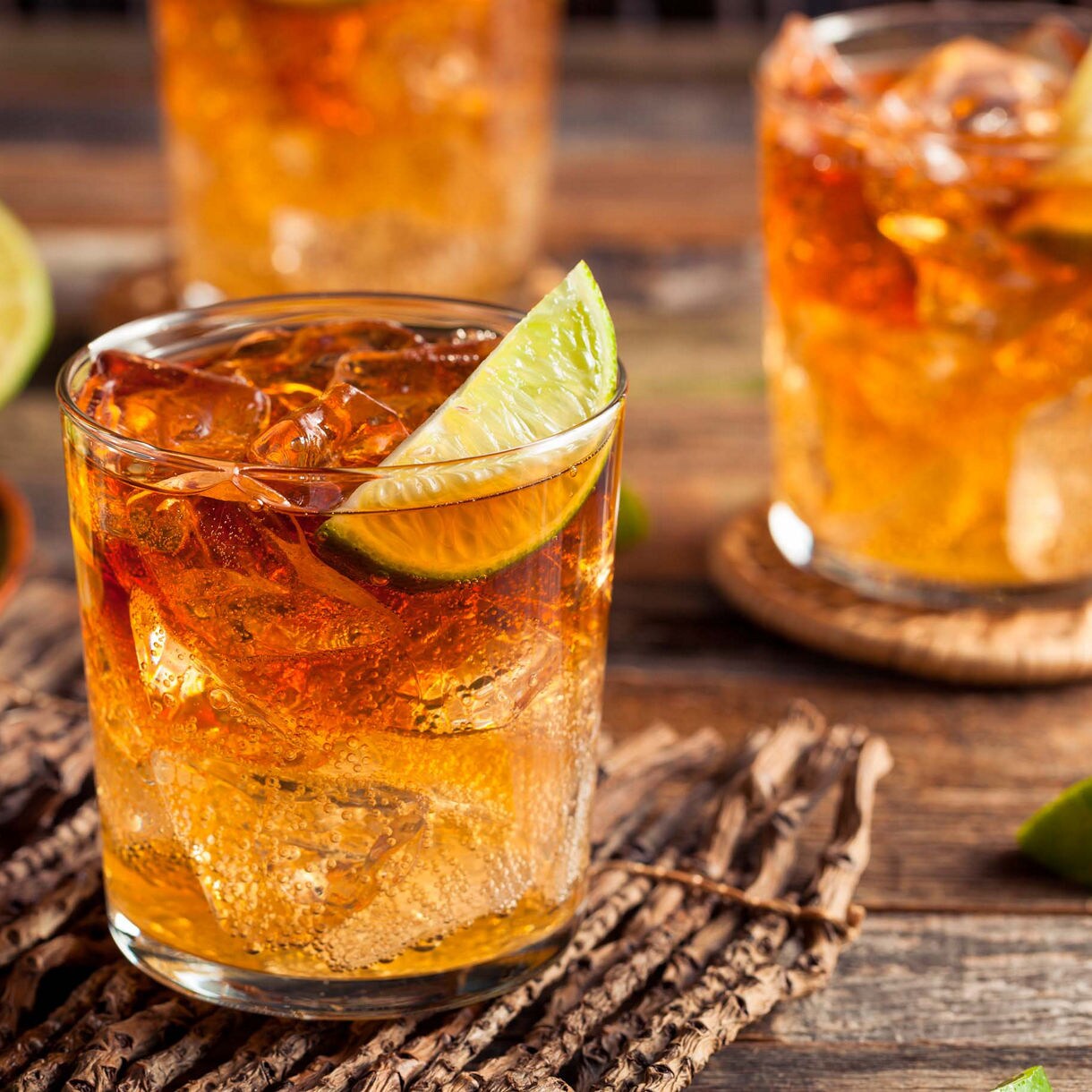 A glass of iced cocktail featuring a lime wedge garnish sits on a rustic wooden surface. Fresh lime slices are displayed in a bowl nearby, adding a vibrant touch to the setting. The drink's amber hue and condensation evoke a refreshing mood, perfect for casual gatherings or summer evenings.