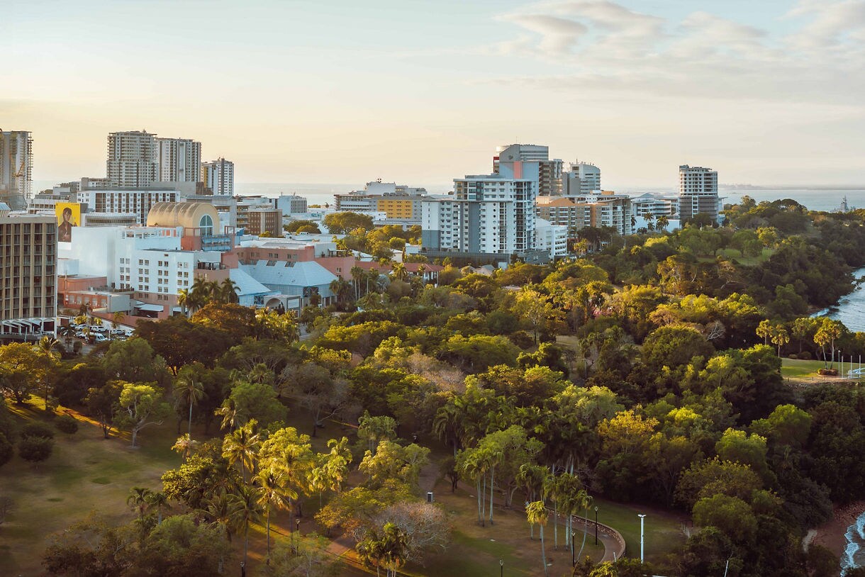 Aerial view of Darwin’s waterfront skyline at sunset, showing modern buildings, lush parkland and the blue waters of the Timor Sea.