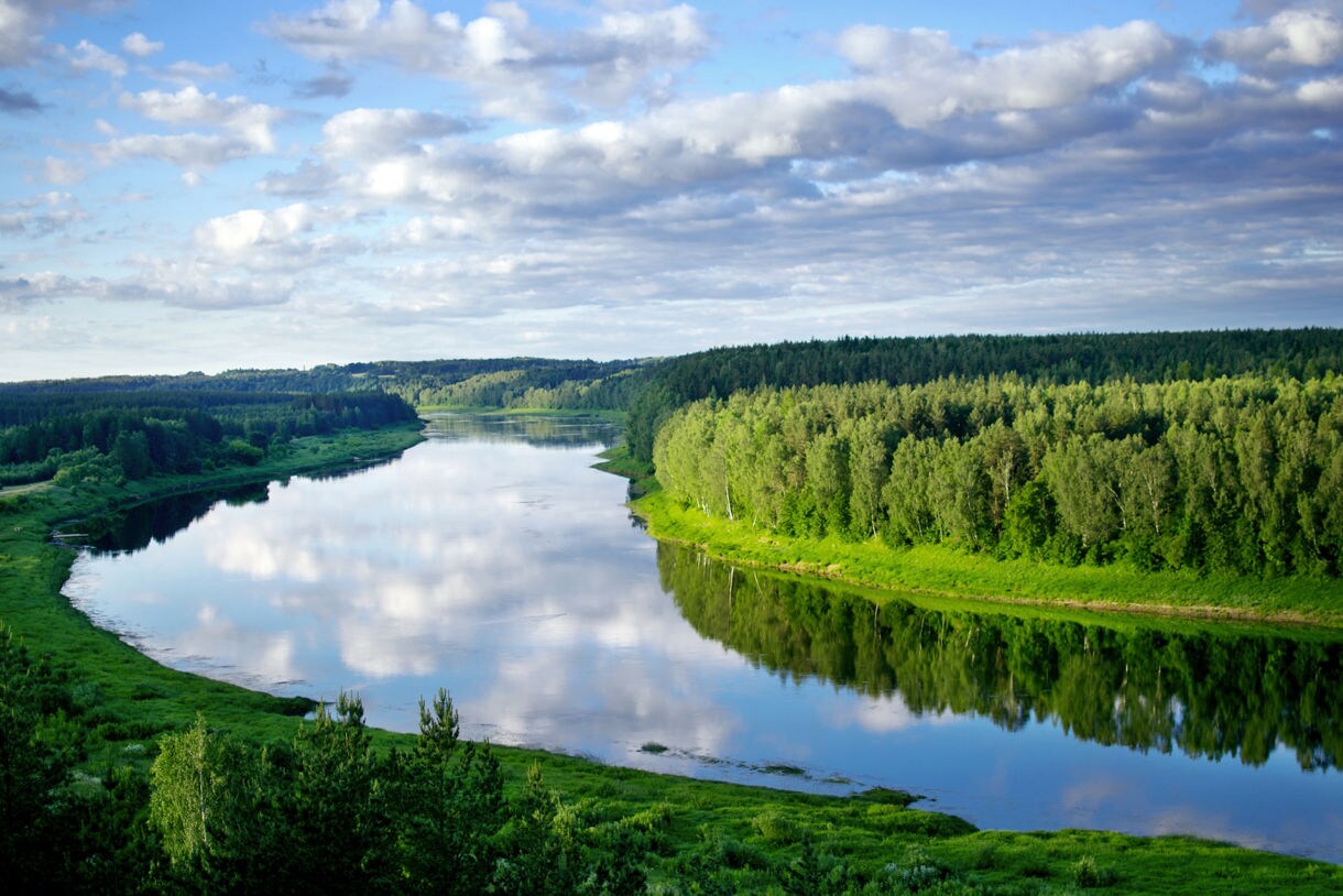 Scenic view of the Daugava River in Latvia, reflecting blue skies and clouds, surrounded by dense green woodland and grassy banks.