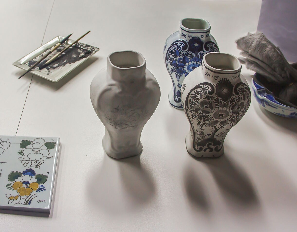 A tabletop scene showing three vases at different stages of Delft pottery painting, surrounded by brushes, paint trays and a sample tile with floral designs.