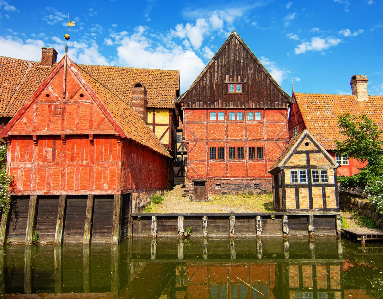 Alt: Historic half-timbered houses with red and yellow walls reflected in a canal at Den Gamle By in Aarhus, Denmark.