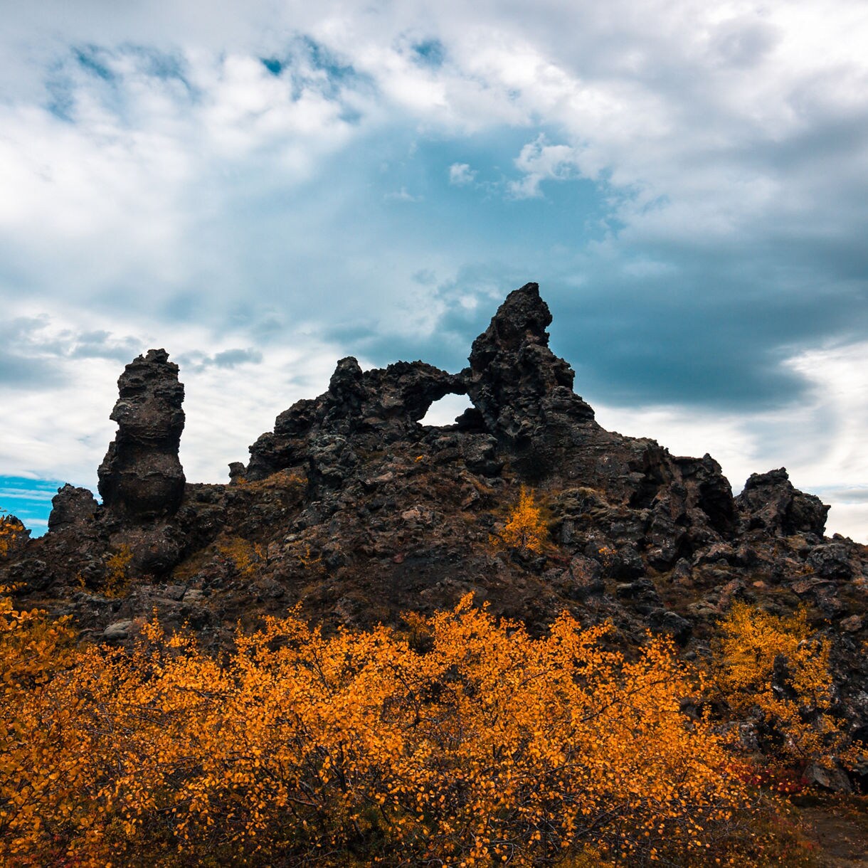 Dark volcanic rock formations at Dimmuborgir in Iceland, with tall lava pillars and a natural arch framed by vibrant yellow autumn trees under a dramatic cloudy sky.