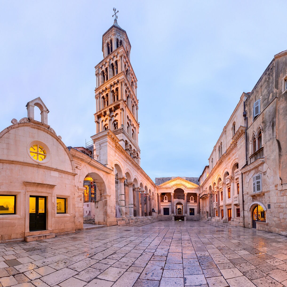 Open courtyard in Split with the tall bell tower of the Cathedral of Saint Domnius, arched colonnades and surrounding stone buildings glowing softly at dusk.