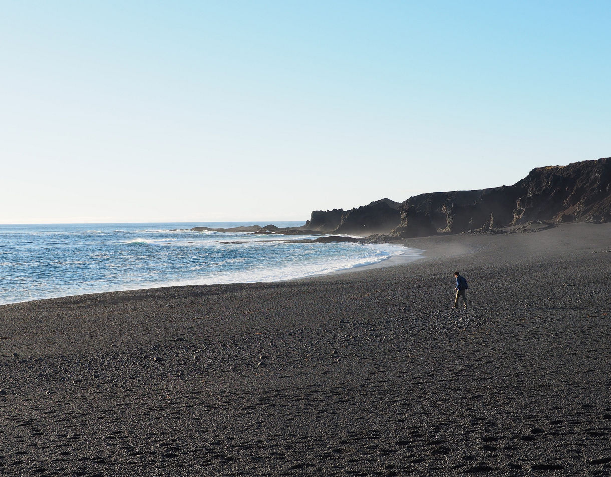 A lone person walking on a wide black pebble beach with gentle ocean waves and jagged dark cliffs in the distance under a clear sky.