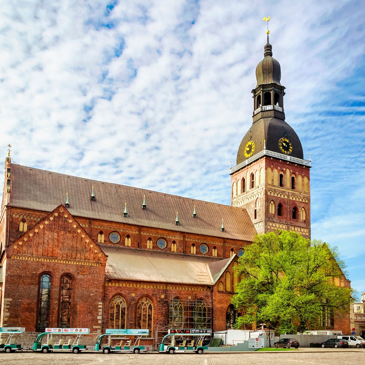 Exterior view of Riga Dome Cathedral in Latvia, featuring a large brick façade with Gothic windows and a tall black-domed clock tower rising against a partly cloudy blue sky.