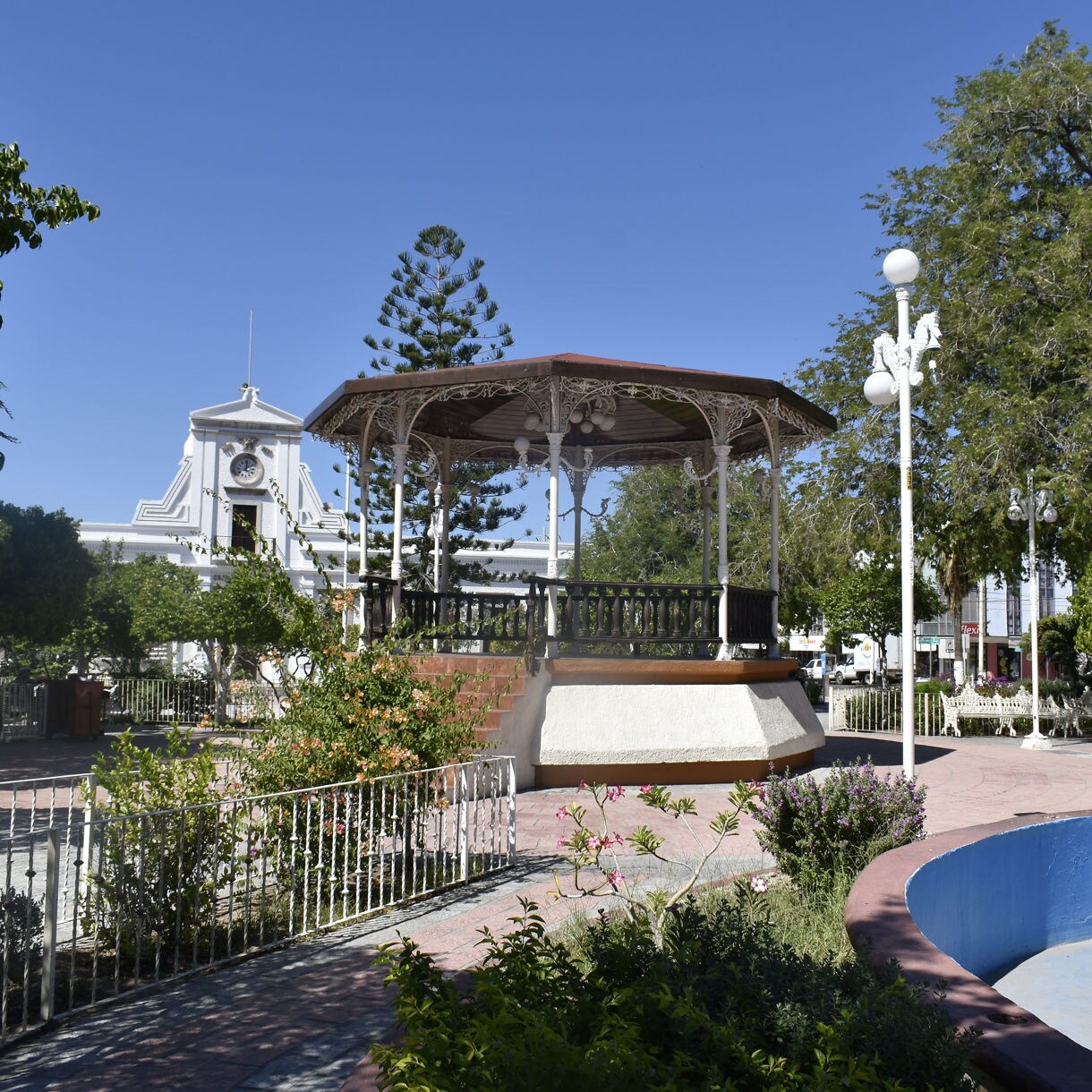 Park in downtown La Paz featuring a central gazebo, clock tower building in the background, trees, garden plants and a circular fountain.