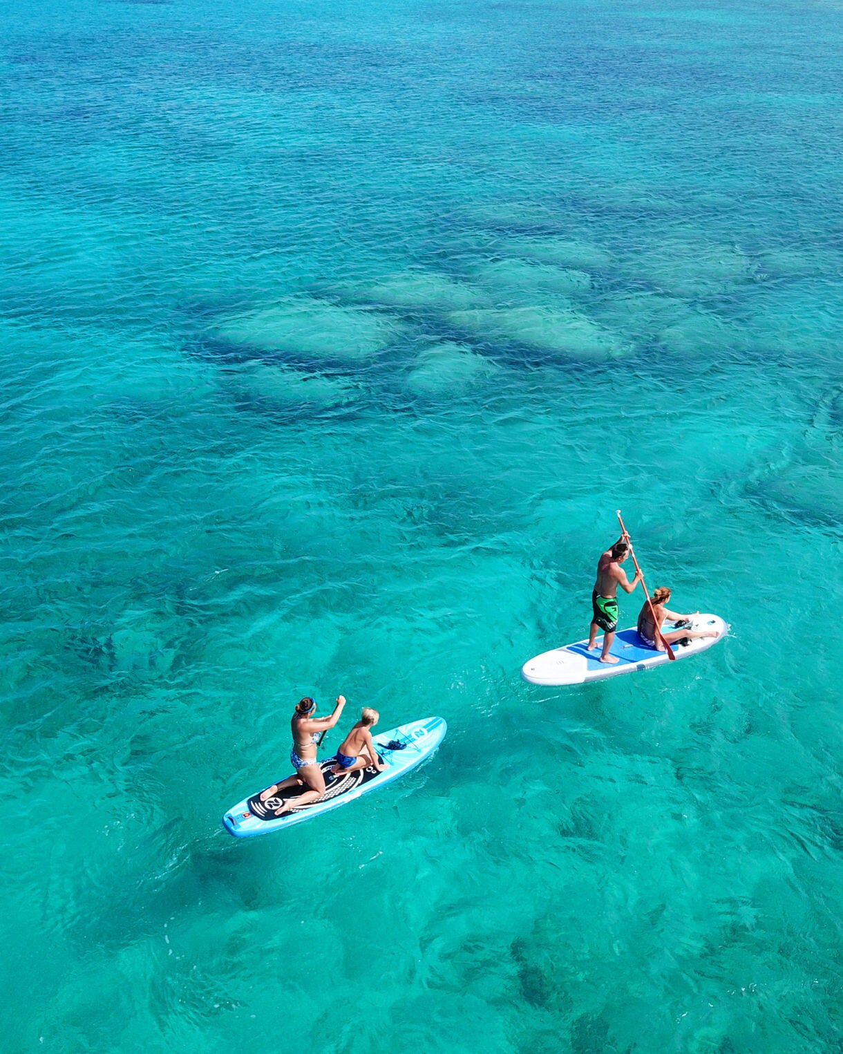 Two people on paddleboards gliding over calm, turquoise water near a sandy shoreline on Dravuni Island under a bright sky.