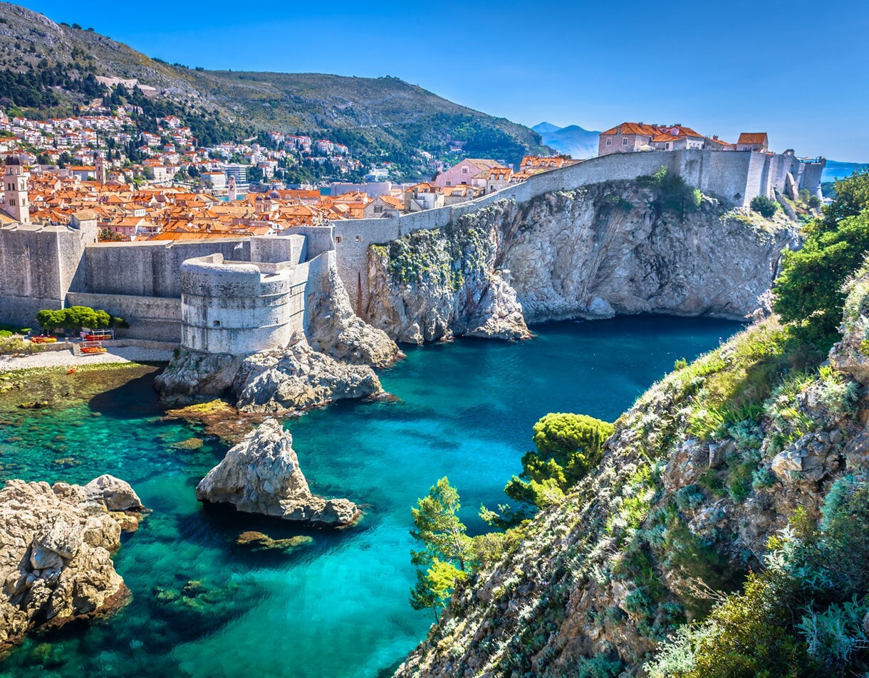 Aerial view of Dubrovnik, Croatia, with medieval stone walls and towers surrounding the old town, red-roofed buildings and cliffs rising above the bright blue Adriatic Sea.