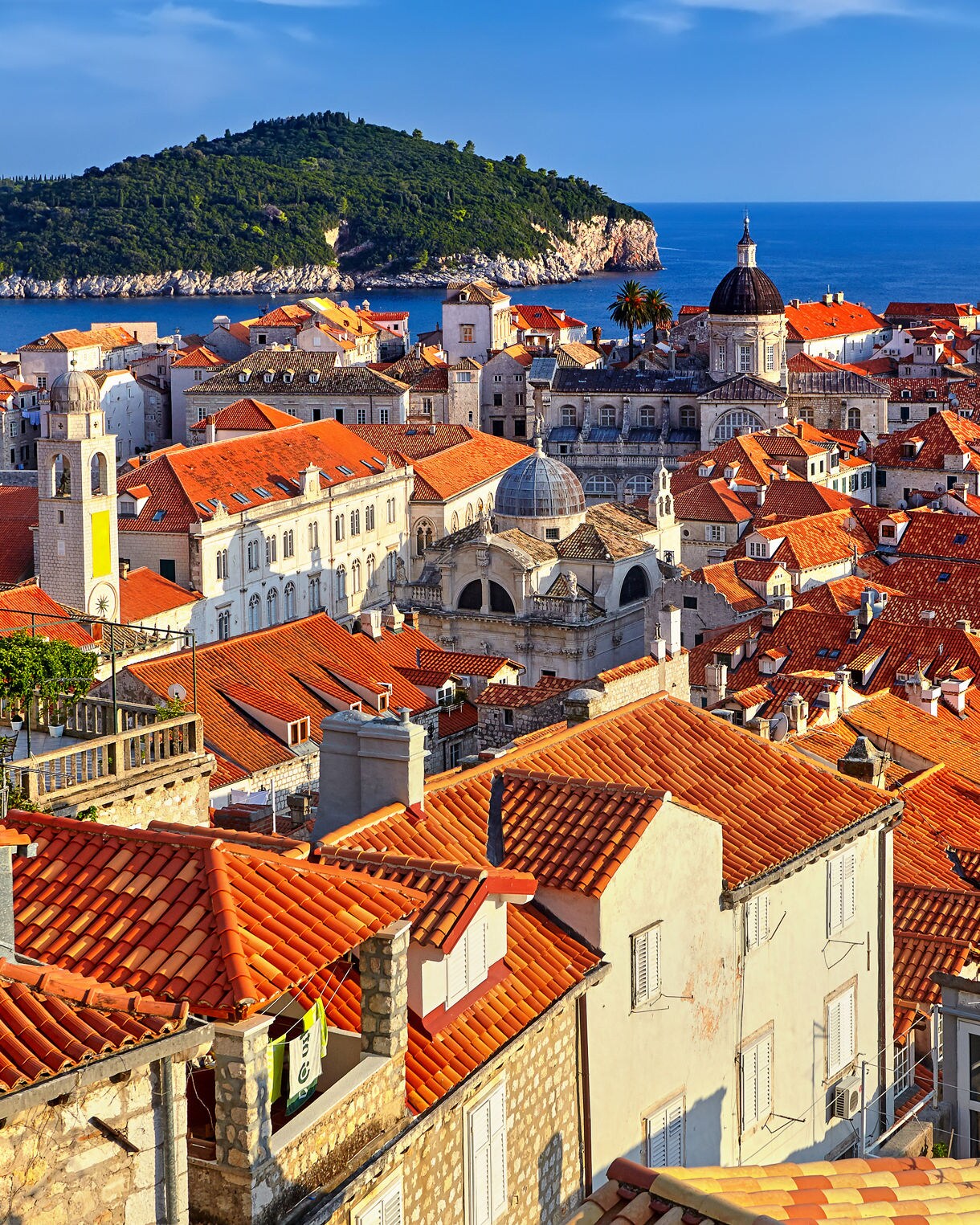Aerial view of Dubrovnik’s Old Town, featuring red-tiled rooftops, historic stone buildings and the sea with a green island in the distance.